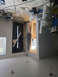 Fotografi av et rom i en bygning som er under renovering eller ombygging. Bildet viser en svart inngangsdør, en åpen dør til et annet rom, en vindu, og diverse byggematerialer og utstyr på gulvet.