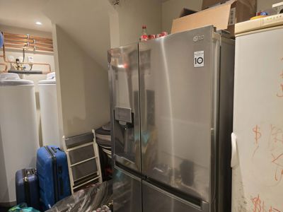 Fotografi av et rom inne i en bygning, sannsynligvis et kjellerrom eller teknisk rom, som viser en stor kjøleskap, varmtvannstanker og rør.