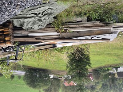 Fotografi av en byggeplass eller et område med materialer (treplater, tømmer, teltduk) i forgrunnen og en landsby i bakgrunnen.