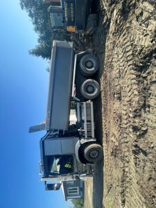 Fotografi av en grå lastebil (kjettingbil) og en gullig graver på en byggeplass med en ferdig hus i bakgrunnen.