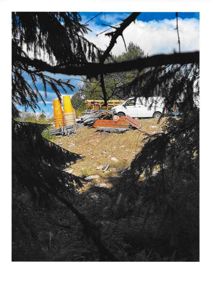 Fotografi av et byggeområde sett gjennom trær, med en hvit varebil og byggematerialer i bakgrunnen.