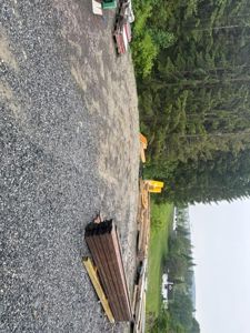 Fotografi av et byggeplassområde med grus, oppstakkede byggematerialer (tre, stål) og skog i bakgrunnen.