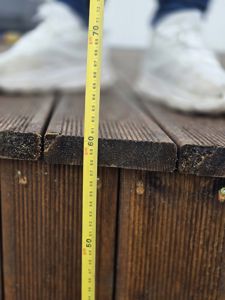 Fotografi som viser en gul målebånd som holder en høyde på 70 cm på en mørk, treliknende overflate (sannsynligvis en terrasse eller dekk). I bakgrunnen er det uskarpe bilder av hvite sko.