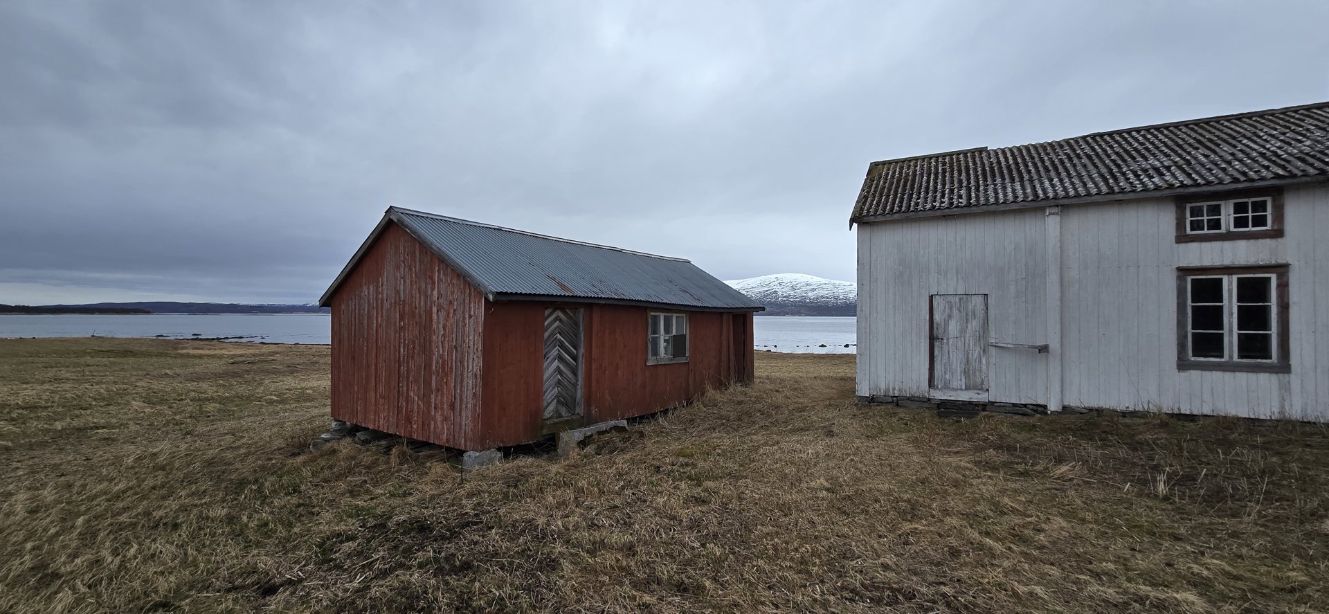 Fotografi av to små, enkle bygninger (en rød og en hvit) på en åpen, gråaktig mark ved kysten. Bildet viser bygningenes utseende og plassering i forhold til hverandre og omgivelsene.