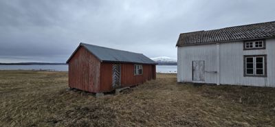Fotografi av to små, enkle bygninger (en rød og en hvit) på en åpen, gråaktig mark ved kysten. Bildet viser bygningenes utseende og plassering i forhold til hverandre og omgivelsene.