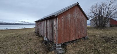 Fotografi av en rød, gammel bygning (kanskje en uthus eller båthavn) sett fra siden, med en snødekket fjellkjede i bakgrunnen.