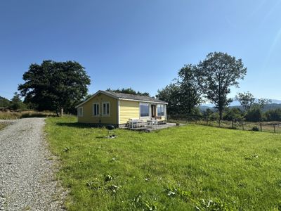 Fotografi av en gul hytte sett fra utsiden, med omgivelser som grøft, trær og en grusvei.