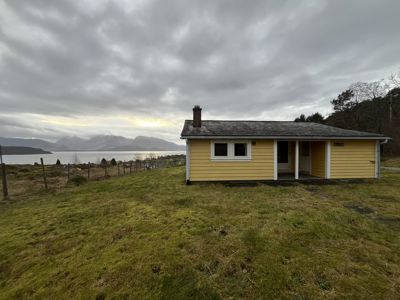 Fotografi av en gul hytte med tak og skorstein, sett fra siden mot et vann og fjell i bakgrunnen.