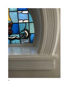 Fotografi av en detalj på en vindusramme, spesifikt et hvitt, profilert vindusomslag (karm) og en del av et farget glasspanel (mosaikkglass) i bakgrunnen.