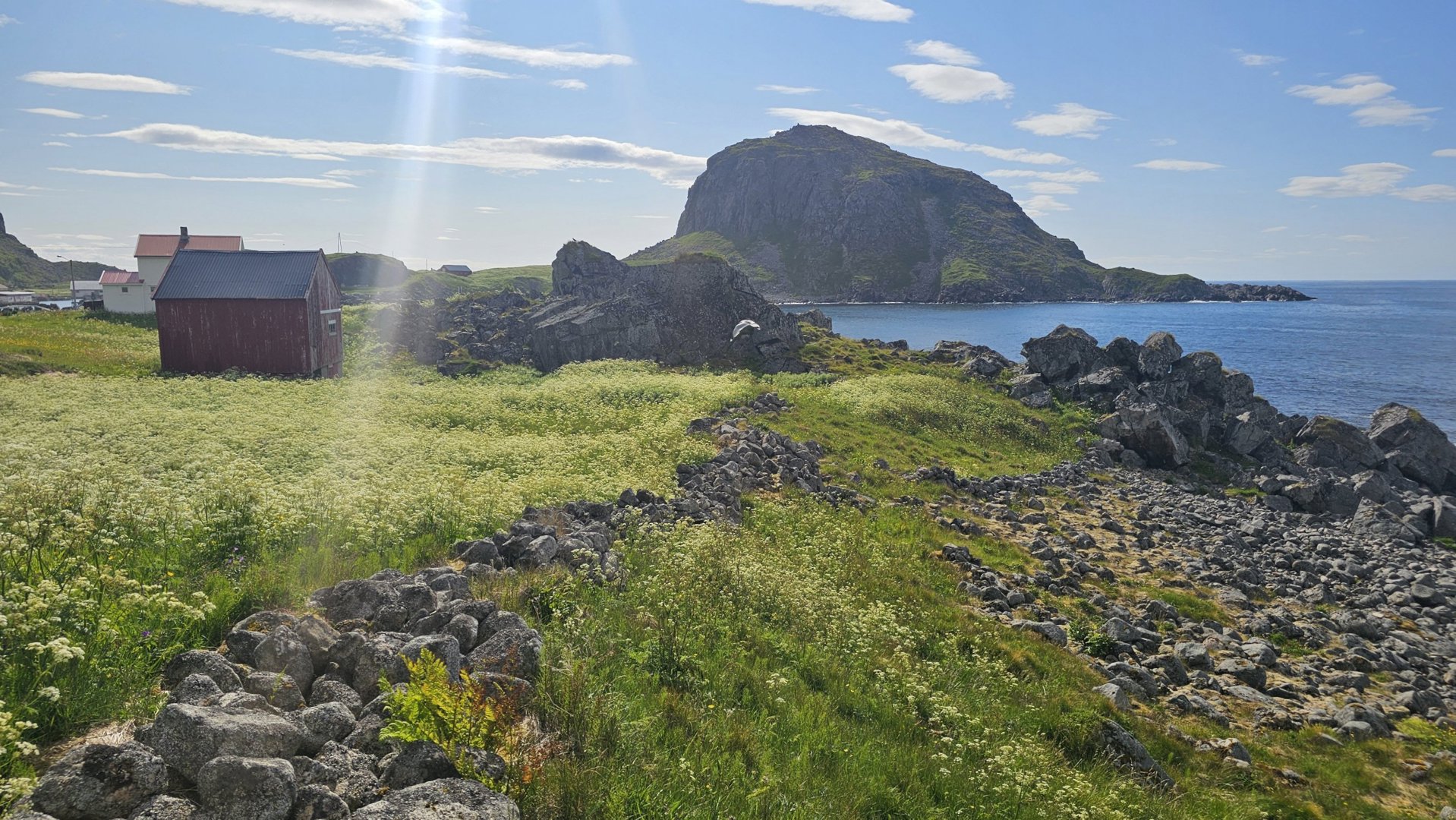 Fotografi av en kystnær bygning (rød uthus) med en stor klippeformasjon og hav i bakgrunnen.