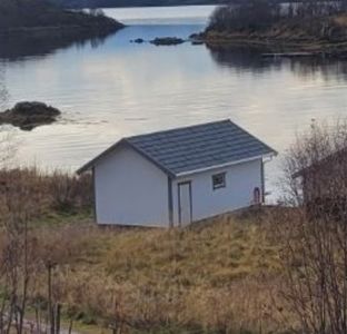 Fotografi av en liten hvit hytte eller uthus plassert på en grøntbelte ved siden av et vann. Bildet viser bygningens fasade og omgivelsene.