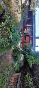 Fotografi av en rød hytte sett fra siden, med omgivende natur, store steiner og en ildstedshyll.