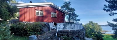 Fotografi av en rød villa med hvit veranda, plassert på en steinete bakke med utsikt over vann.