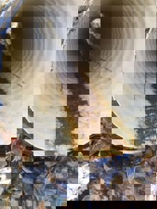 Fotografi av en stor, hvit, ribbet rørledning (sannsynligvis en avløpsrør eller vannledning) sett innfra. Røret er delvis fylt med mørk, slammete vann og sediment. Bildet viser også omgivelsene med fuktig bakke, løv og steiner.