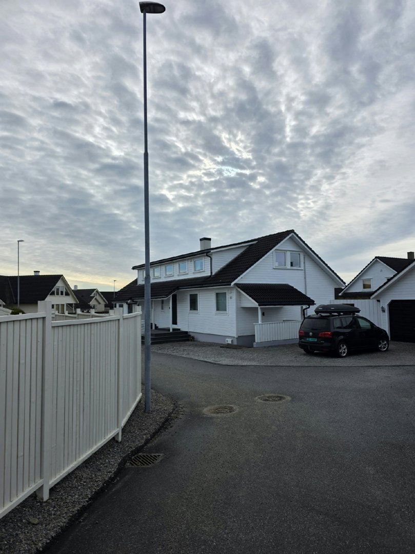 Fotografi av en hvit villa med mørk takflate, sett fra en vei med en hvit hekk i forgrunnen.