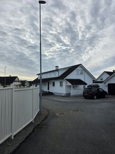 Fotografi av en hvit villa med mørk takflate, sett fra en vei med en hvit hekk i forgrunnen.