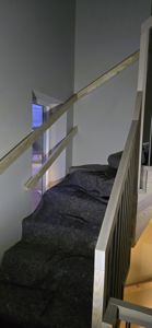 Fotografi av en trapp i en bygning under ombygging eller renovering. Trappetrinnene er dekket med mørk beskyttelsesplast, og det er installert midlertidige håndtak i ufinert tre.