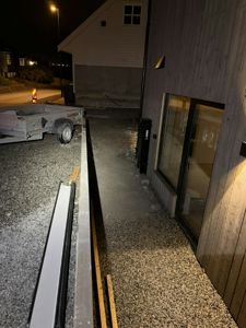 Fotografi av en bygning under eller etter utvendig arbeid, tatt om natten. Bildet viser fasaden, en stor glassdør, en kjørevei med grus, og en tilhenger i bakgrunnen.