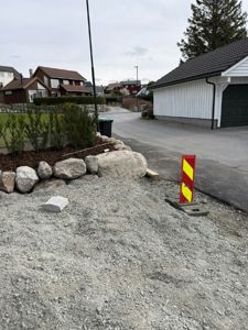 Fotografi av en byggeplass eller tomt i et boligområde, med grus, steiner, en garasje og nabolagshus i bakgrunnen.