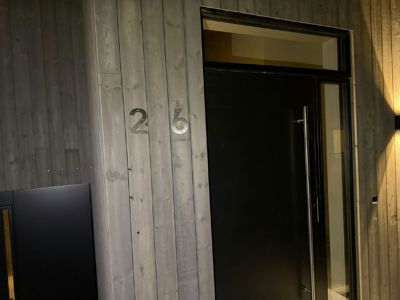 Fotografi av en bygningsside med en mørk dør, vindue og et husnummer (26) på en trepanelvegg.