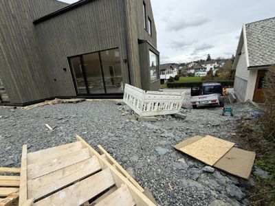 Fotografi av en byggeplass med en ferdig fasade på en nybygget bolig, grus under, og byggemateriale i forgrunnen.