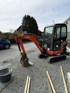 Fotografi av en liten graver (Kubota) på en grusflate, med en bil og hus i bakgrunnen.