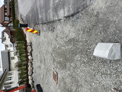 Fotografi av en grusbelagt vei eller parkeringsplass med en steinmur, et avløpsrør og et gult og røtt skilt i forgrunnen. I bakgrunnen er det flere bolighus.