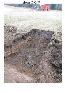 Fotografi av en utgravet grøft eller fundamenteringsgrøft på et byggefelt, med en rød skur i bakgrunnen.