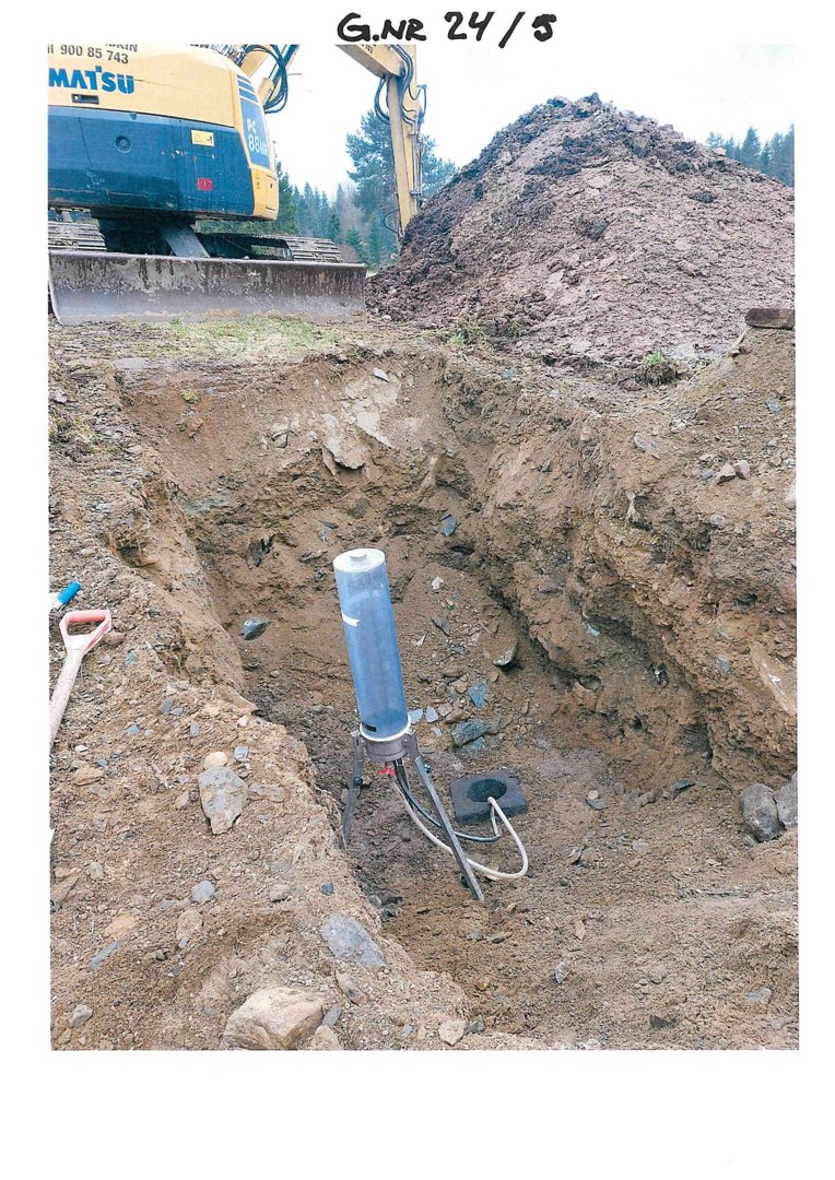 Fotografi av en utgravet grøft eller hull på et byggeplass, med en gaffeltruck i bakgrunnen og et måleinstrument (sannsynligvis for grunnvannsmåling) plassert i grøften.