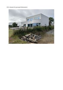 Fotografi av en moderne, hvit villa sett fra siden. Bildet viser bygningens fasade, omgivelsene med grusvei, steiner og vegetasjon, samt en del av en nabobygning i bakgrunnen.