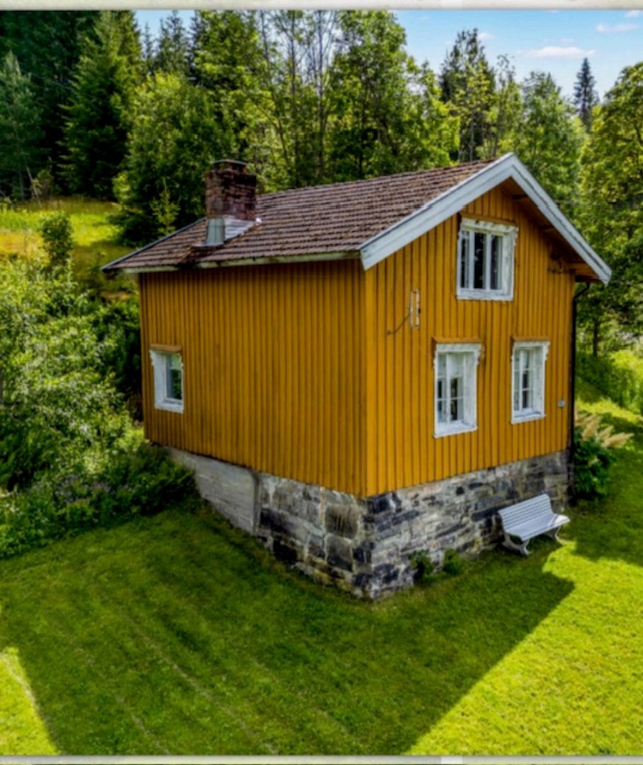 Fotografi av en gul, trepanelt hytte med steinfundament, sett fra en vinkel som viser både fasade og sidevegg. Hytta er plassert på en grønn hage med skog i bakgrunnen.