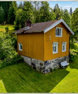 Fotografi av en gul, trepanelt hytte med steinfundament, sett fra en vinkel som viser både fasade og sidevegg. Hytta er plassert på en grønn hage med skog i bakgrunnen.