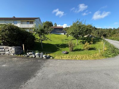 Fotografi av en eksisterende bygning (hus) sett fra en vei, med omgivelser som hage, trær og skog.