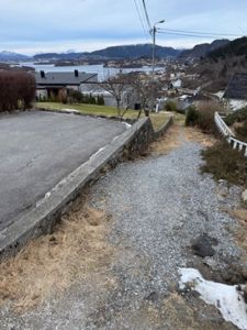 Fotografi av en tomt med en betongkjørevei, en grusvei, en steinmur og nabolag med fjord og fjell i bakgrunnen.