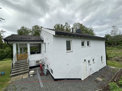 Fotografi av en hvit, toetasjes bolig sett fra siden. Bildet viser fasaden, vinduer, tak, trapp opp til en veranda og omgivelser med grus og gress.