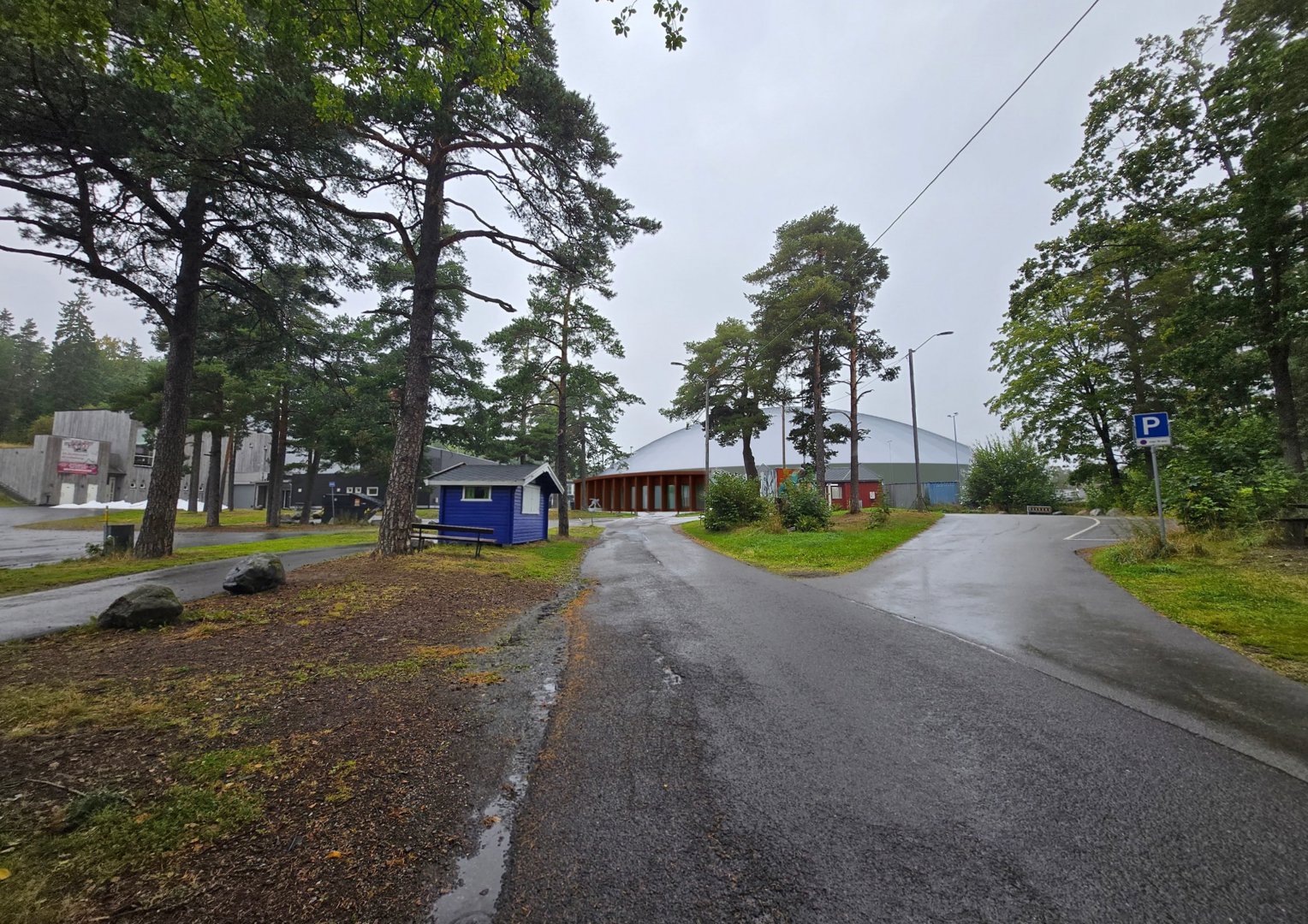 Fotografi av en bygning (en stor hall med hvit tak) sett fra en vei, omgitt av trær og en liten blå hytte.