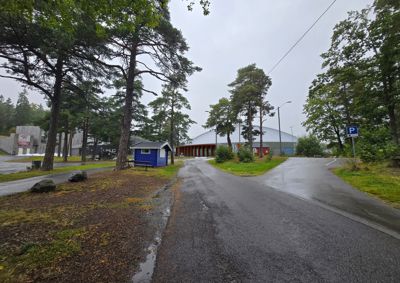 Fotografi av en bygning (en stor hall med hvit tak) sett fra en vei, omgitt av trær og en liten blå hytte.