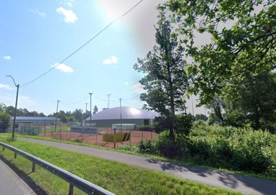 Fotografi av en bygning med hvit, kuppelformet tak (muligens en tennisbane eller lignende) sett fra en vei, med omgivende natur og infrastruktur.