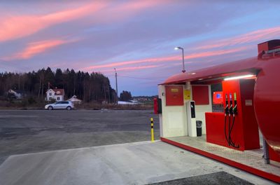 Fotografi av en bensinstasjon med røde bensinpumper, sett mot en himmel med rosa og blå farger ved solnedgang. I bakgrunnen er det en skog og en hvit bil.
