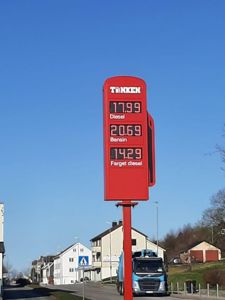 Fotografi av en bensinstasjon med prisliste for diesel, bensin og farget diesel, med hus og en lastebil i bakgrunnen.