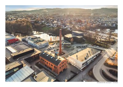 Et fotografi (flyfoto) som viser en eksisterende situasjon av et område med flere bygninger, inkludert en stor rødt teglbygning og en høy skorstein. Bildet er merket 'Eksisterende situasjon - nordøst'.
