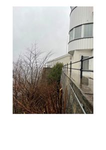 Fotografi av en hvit bygning med rund hjørne, sett fra siden med en steinmur og gjerde i forgrunnen.