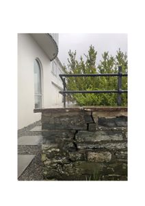 Fotografi av en steinmur med en svart metallrailing foran, sett fra siden. I bakgrunnen ser man en hvit fasade med et buet vindu og en del av taket.