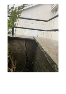 Fotografi av en betongkonstruksjon (kanskje en trapp eller kant) med en tydelig, vertikal sprekk i betongen. Bakgrunnen viser en hvit fasade og en metallrailing.