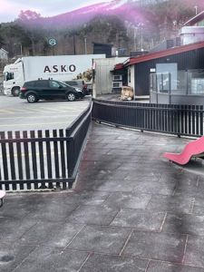 Fotografi av en utendørs plass med asfaltert gulv, svart hekk og et barneslideo i forgrunnen. I bakgrunnen er det parkerte biler (en lastebil med 'ASKO' logo) og en bygning.