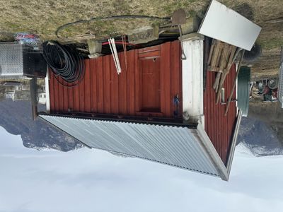 Fotografi av en liten, rød skur eller uthus med hvit taklist og metalltak, sett fra siden. Bakgrunnen viser fjell og en del av en annen bygning.