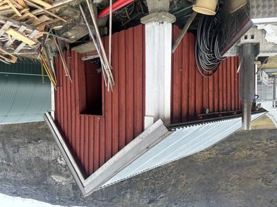 Fotografi av en liten, rød bygning (trolltun eller uthus) med hvit taklist og hvit malte søyler. Bildet viser også omgivelser med byggematerialer og en grønn bølgeplatevegg i bakgrunnen.