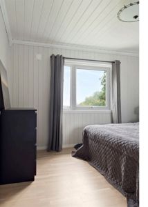 Fotografi av et soverom med hvite panelvegger, gulv, vindu og møbler.