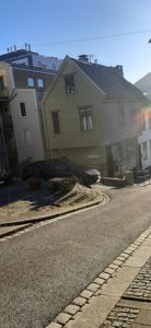 Fotografi av en gulett trehus med skrå tak, parkert bil og nabohus på en gate.