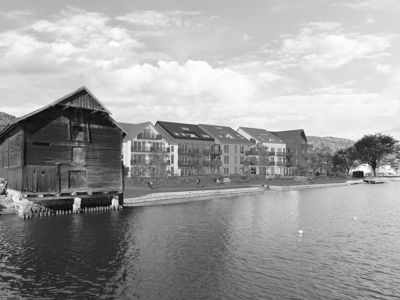 Fotografi som viser en eksisterende bygning (en gammel tømmerbygning) ved vannet, med nye boliger i bakgrunnen.